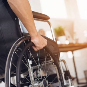 A disabled man is sitting in a wheelchair. He holds his hands on the wheel. Nearby are his colleagues